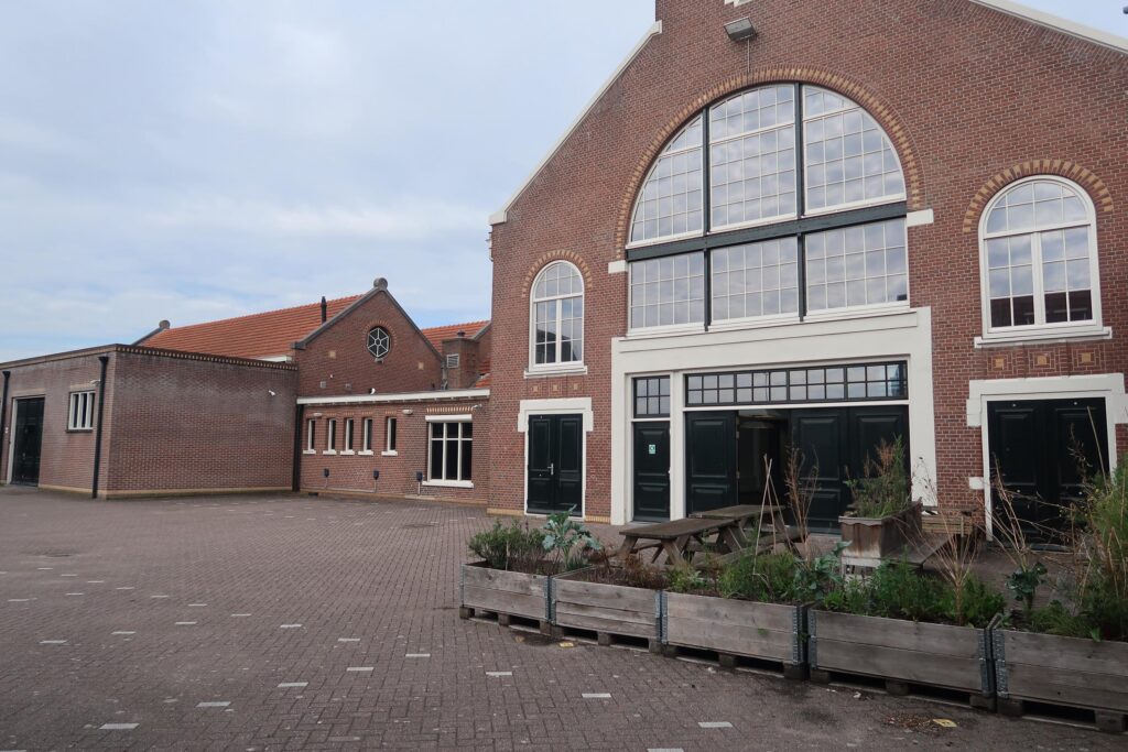 Vooraanzicht van het voormalige fabriekspand aan het Gedempt Hamerkanaal met grote ramen, zwarte deuren en een betegeld plein met plantenbakken.